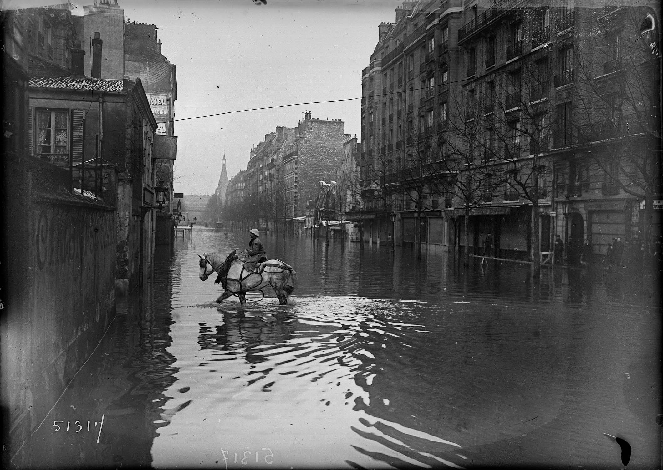 Cavalière en amazone traversant à cheval une rue inondée ; enseignes et publicités
