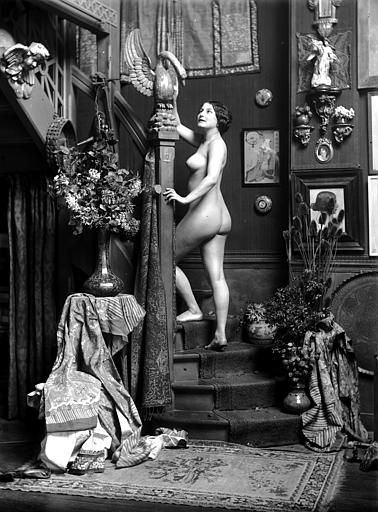 Mise en scène d'un modèle féminin : jeune femme nue montant un escalier, vue de trois-quart dos, statue de cygne posée sur le montant de la rampe d'escalier