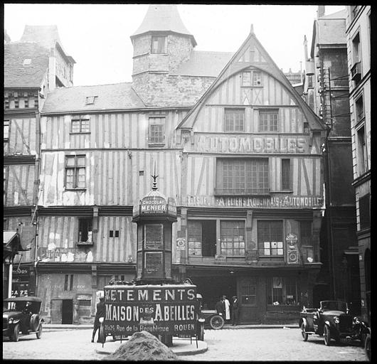 Maisons à pans de bois et garage automobile ; au premier plan, vespasienne couverte d'inscriptions publicitaires