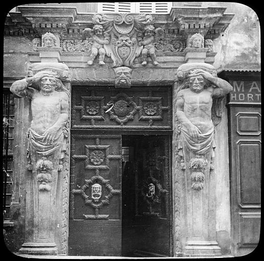Porte ancienne  flanquée de deux atlantes sculptés ; au centre du linteau, putti encadrant un blason