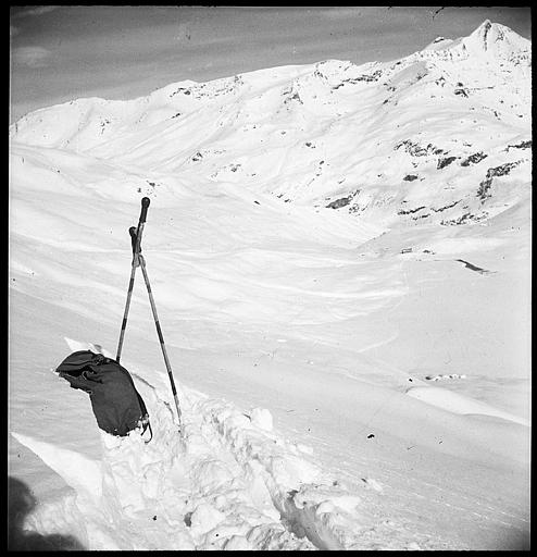 Dans la neige, au premier plan, un sac et deux bâtons de ski