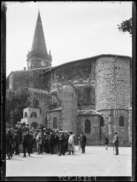 Vue partielle du chevet avec l’absidiole encore surmontée par sa tour de défense du 16e siècle. Groupe de promeneurs autour du calvaire.