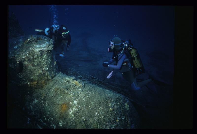 Vue sous-marine de deux plongeurs face à la carène métallique.
