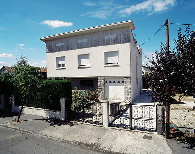 Vue d'ensemble ; un garage occupe le côté droit du rez-de-chaussée, tandis qu'à gauche des pilotis supportent le 1er étage, sous un toit en dévers.