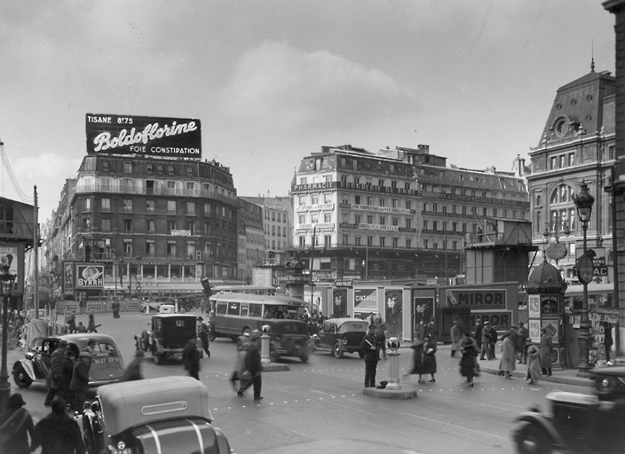 Vue de la place devant la gare Saint-Lazare. Un des immeubles est surmonté d'une grande enseigne lumineuse pour la Boldoflorine.