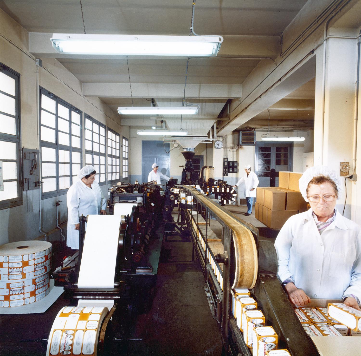 La chaine de conditionnement du 2e étage du bâtiment principal en activité. A gauche, la machine fabriquant des boites pour le vrac (à partir de papier cartonné imprimé et de papier fin formant le sac intérieur). Les boites étaient ensuite remplies de tisane à l'aide de la machine au fond (sous le grand entonnoir) et revenaient sur un tapis roulant (le temps que la colle sèche) pour être stockées dans des cartons d'expédition.