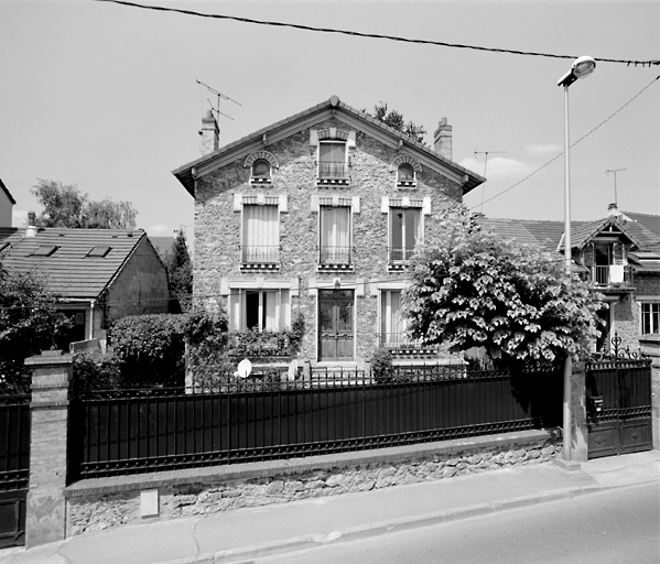 Vue d'ensemble de la maison. Elle s'abrite derrière une grille doublée de plaques de tôle.