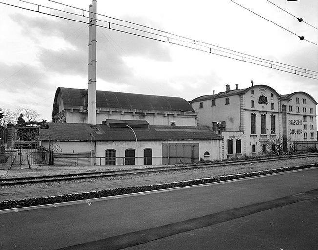 Vue d'ensemble, depuis la voie ferrée au nord : partie ouest de la brasserie Gruber. On voit, de gauche à droite : la passerelle ferroviaire, la chaufferie (et derrière, le cuvage), la salle à brasser et les silos.