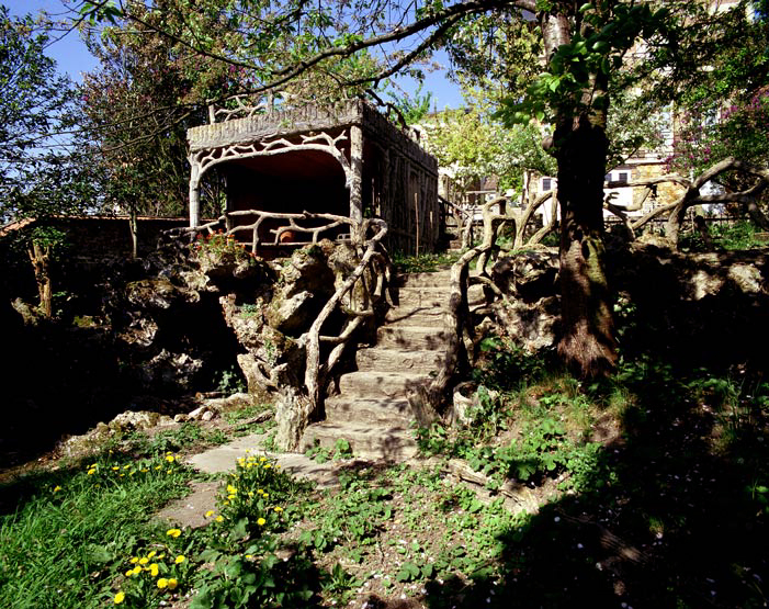 Vue des constructions en faux bois écoté qui ornent le jardin (kiosque et escalier). Entre le kiosque et l'escalier, une grotte de rocaille abrite une source.