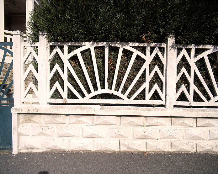 Clôture en ciment. Les panneaux ajourés reposent sur un muret en parpaings moulés ; le motif de ces panneaux est repris en métal à la porte d'entrée.