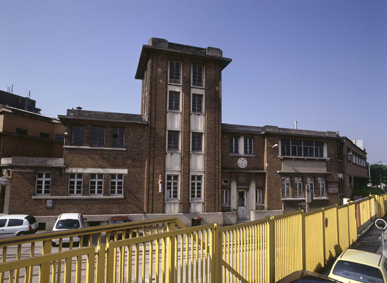 Façade de la 'boulangerie' (fournil et moulin d'essai). Construite en 1933, parallèlement aux voies de chemin de fer, et marquant ainsi l'entrée du site, ce bâtiment offre un corps central à trois étages carrés coiffé d'une corniche en béton et d'un toit-terrasse ; la façade en parement de brique, dissimulant une structure en béton armé, porte le monogramme ' GMPP ' (Grands Moulins de Pantin-Paris).