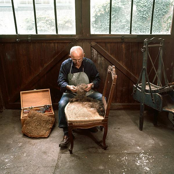 M. *, qui a fait son apprentissage dans l'atelier de tapisserie, restitue quelques gestes du travail. Après le garnissage du siège, la mise en crin (précédant la piqûre crin) permet d'uniformiser le fond et d'éliminer les bosses avant la mise en blanc, la pose d'une feuille de ouate et la couverture.