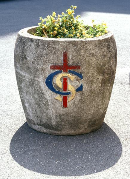 Jardinière en ciment dans le parc de l'hôpital portant la croix et les initiales de saint Camille entrelacés.