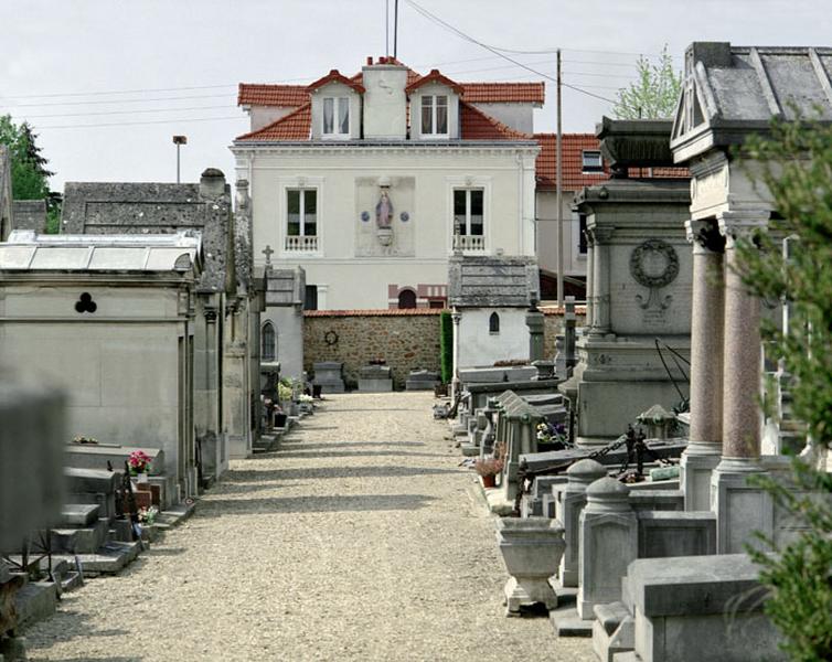 Vue d'ensemble d'une allée vers la rue Louis Delamarre. Dans l'axe de la première allée, se trouve une maison fermant la perspective par une niche ornée d'une Vierge sculptée. Cet édifice était la première maison du conservateur du cimetière.