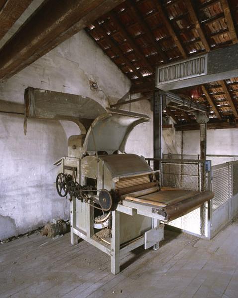 Vue du mécanisme de fonctionnement d'une cardeuse à un seul tambour cardant située à l'étage de combles de l'entrepôt industriel. Au second plan, monte-charge de l'entrepôt.