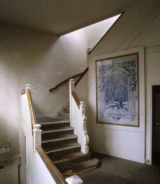 Vue intérieure de l'escalier en bois à départ de rampe sculpté situé dans le bâtiment administratif. Sur le mur d'échiffre, un panneau de céramique à décor de grisaille.