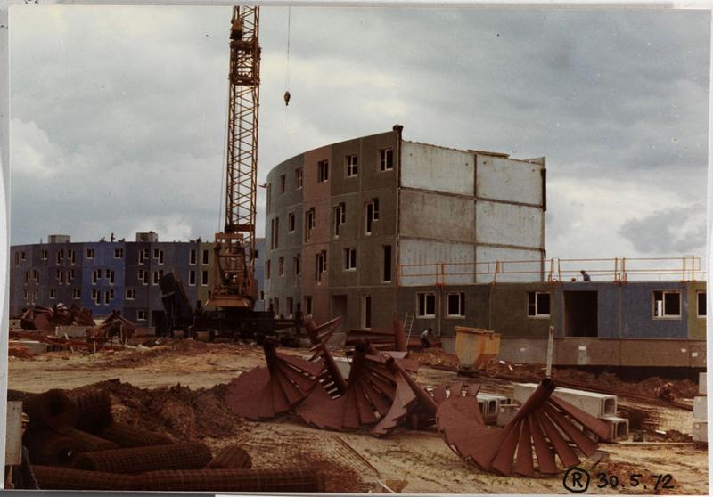 Vue du chantier, bâtiment courbe en construction au second plan ; au premier plan, escaliers pré-fabriqués stockés au sol.