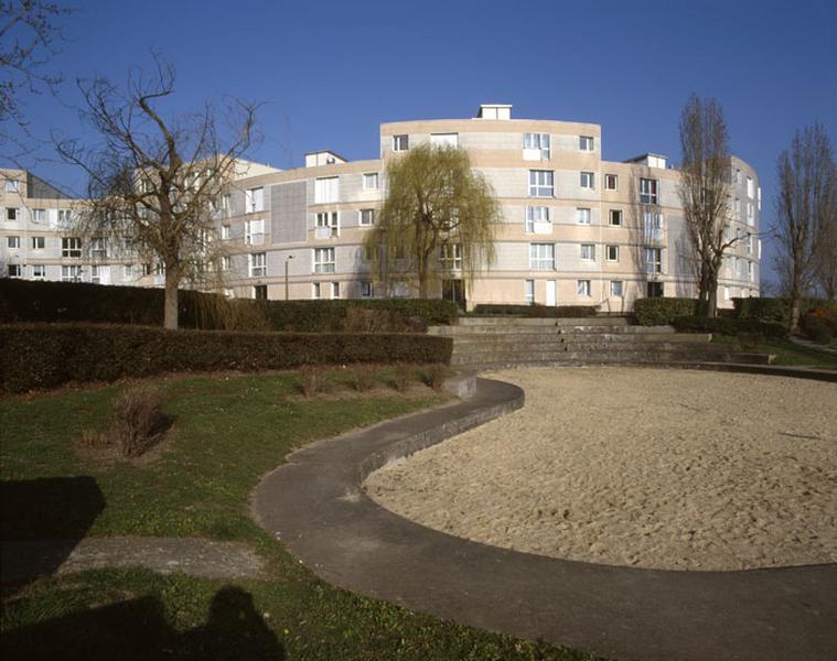 Autre vue actuelle du bâtiment serpentin de l'Ellipse. Au premier plan, une aire de jeu : étang de sable bordé de gradins comme un théâtre.