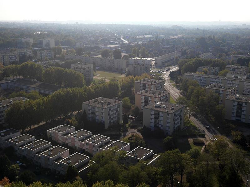Au premier plan, 2 ensembles de logements individuels en bande (situés à l'arrière du centre social des Amandiers) et 4 plots le long de la rue Frédéric-Joliot-Curie (le dernier séparé des 3 autres regroupés autour de la place Edith-Piaf, par la rue Romain-Rolland, non visible sur la photo) ; à gauche, derrière une rangée d'arbres, le groupe scolaire Romain-Rolland.