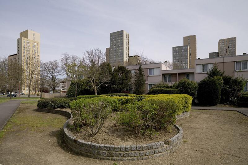 Massif d'arbustes taillés entourés d'une bordure maçonnée devant les logements individuels en bandes situés près de la place Ampère et de la rue Jules-Vallès.