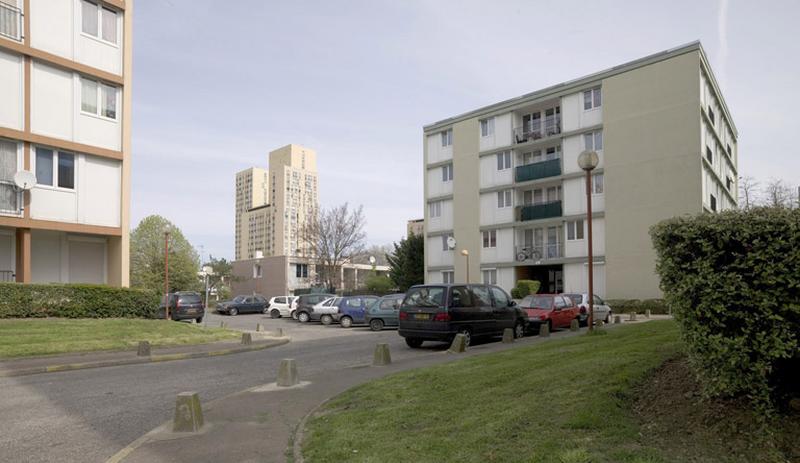 La voie d'accès à la place Ampère entourée d'immeubles cubiques et débouchant sur les bâtiments individuels en bande de l'allée Antoine-Lavoisier.