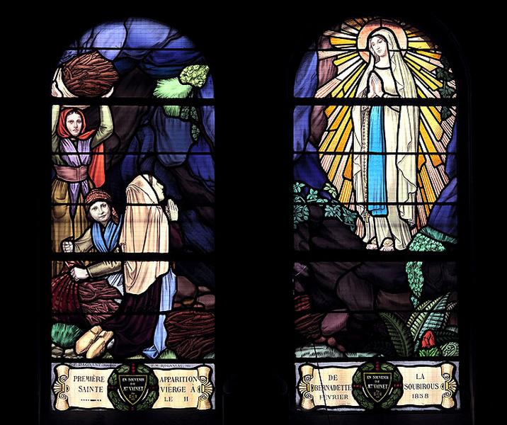 Baie 14 ? :'Première apparition de la vierge à Bernadette Soubirous le 11 février 1858'.La Vierge apparaît à Bernadette ainsi qu'à sa soeur Marie et à une voisine Jeanne Abadie qui ramassait du bois. La Vierge est dans une ouverture du rocher tenant à son bras droit un chapelet avec deux roses posées sur ses pieds ainsi que la scène a été décrite par Bernadette. Oeuvre du peintre verrier Pierre Gustave Dagrant d'après un carton du peintre François Maurice Roganeau.