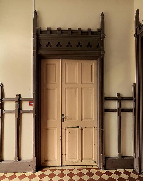 Vue de l'une des portes du palier au rez-de-chaussée. Le caractère néogothique de l'architecture de l'édifice est ici illustré par le traitement du linteau percé de motifs trilobés et surmonté d'un crénelage encadré de deux fleurons.