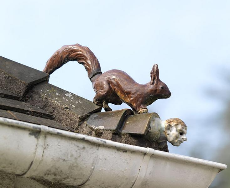 Détail d'un ornement de faîtage (antéfixe) en céramique vernissée, avec représentation animale et humaine : écureuil et visage humain sur l'angle du toit des communs.