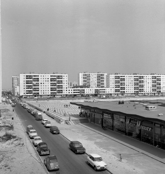 La place de France et le centre commercial. En arrière-plan, les barres de l'ensemble n°3 dit opération Lechauguette. Photographie, 1966. (Ministère de l'Ecologie, MRU 43741027)
