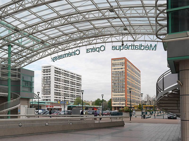 La place de France avec deux des trois barres construites par les architectes Pierre Sonrel et Jean Duthilleul vue depuis le parvis piétonnier de l'opéra. Détail de la toiture du parvis (charpente métallique et plexiglas) laissant passer la lumière. Façades des locaux latéraux réalisées en panneaux d'aluminium gris et vert pistache.