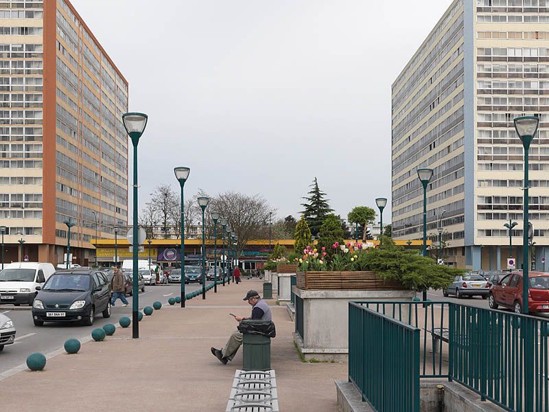 Le parking de la place de France. Au premier plan, accès au parking souterrain surmontés de bacs fleuris. A l'arrière-plan, les commerces en rez-de-chuassée et les deux des barres R+16.