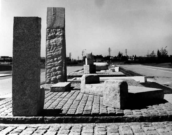 Vue d'éléments de la partie centrale de la composition. Au premier plan à gauche et de dos, «  le siège de l'individualité », à droite « le banc de la collectivité », puis, simple pierre plate « le pont de l'âge adulte », juste derrière, la haute structure de « la porte de la connaissance de soi », au fond, « les cubes de l'enfance » et « l'uf de la conception ». Photographie N/B, [1973].