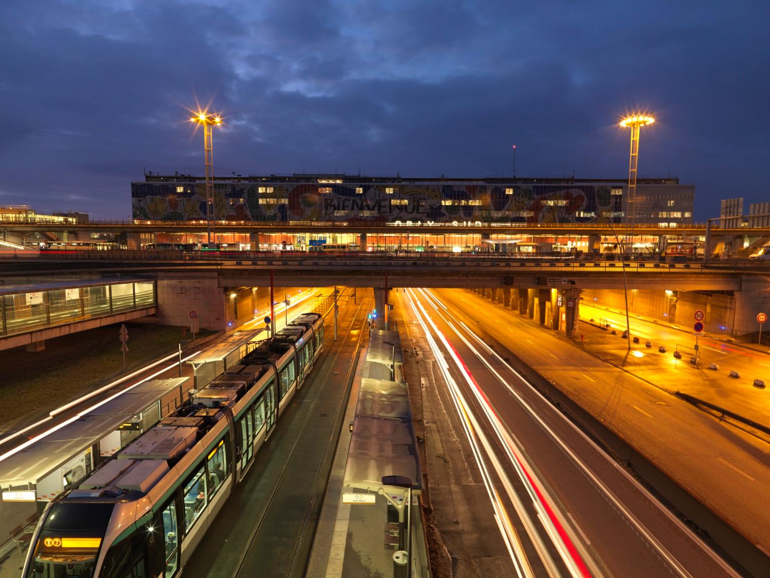 2018, Orly-Sud, la station Aéroport d'Orly du tramway T7 et la Nationale 7, la façade de l'aérogare est décorée d' "Orlove" une œuvre de Jean-Charles de Castelbajac, vue prise depuis le nord. 