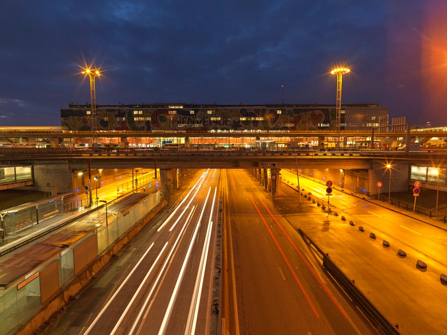 2018, Orly-Sud, la station Aéroport d'Orly du tramway T7 et la Nationale 7, la façade de l'aérogare est décorée d' "Orlove" une œuvre de Jean-Charles de Castelbajac, vue prise depuis le nord. 