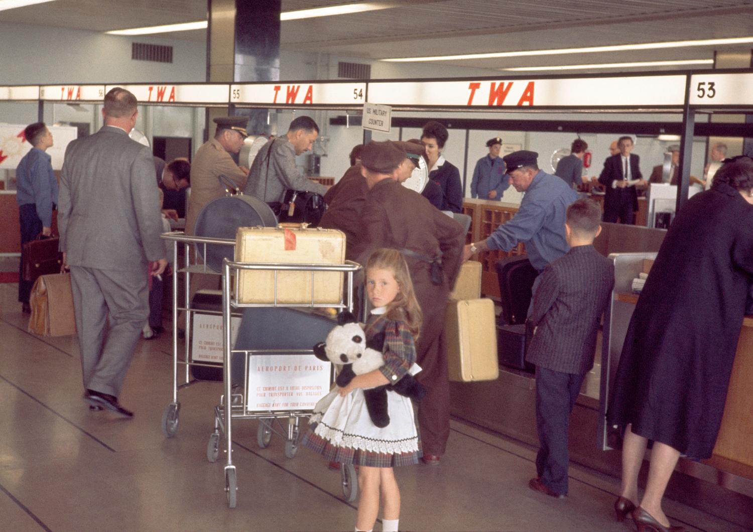 1961, rez-de-chaussée, jeune passagère et son panda devant les comptoirs d'enregistrement de la TWA.