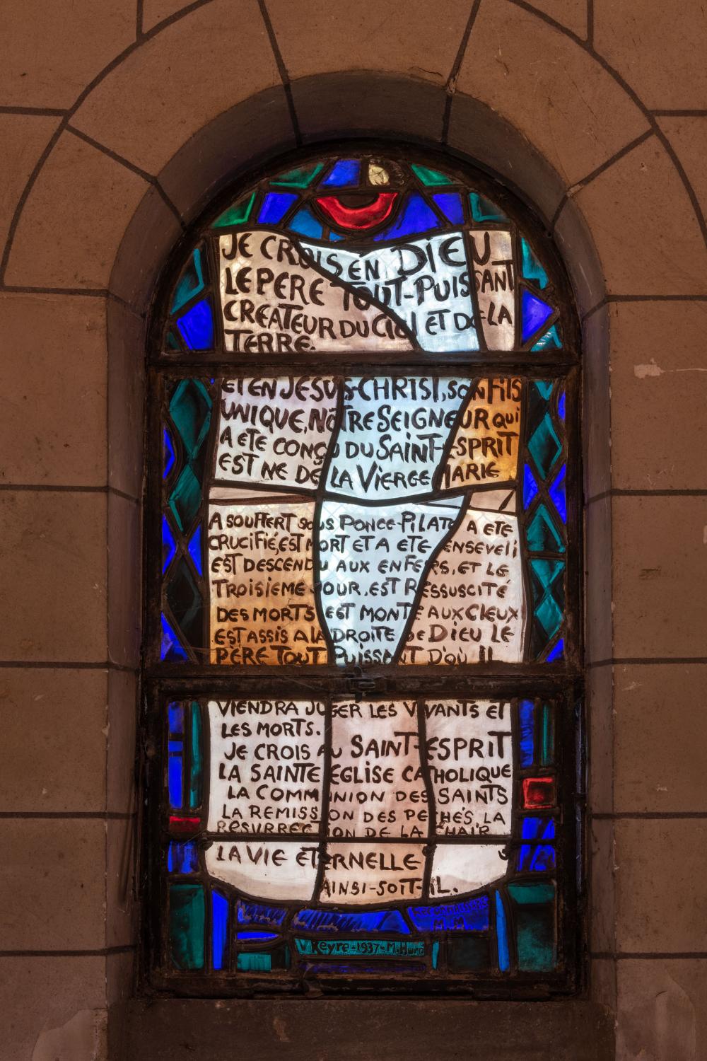 Dans la chapelle-baptistère, vue générale du vitrail de Valentine Reyre représentant le Credo (1937). En bas, la date de 1937 et les signatures de Valentine Reyre et Marguerite Huré. 