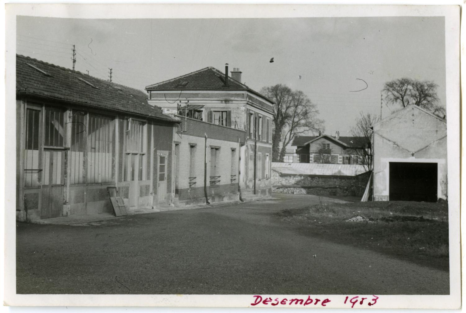 À gauche, les bâtiments du Lysol (murs vitrés et couverture en tuiles mécaniques), puis les bureaux des Ateliers de Technochimie, puis le pavillon d'habitation acheté par les Ateliers de Technochimie en 1959. Au fond, une partie des serres des Grands Etablissements Horticoles Charron Fils. Tirage phot. n & b, 6,5 x 9,5 cm, déc. 1953, sans mention d'auteur (collection Ateliers de Technochimie).