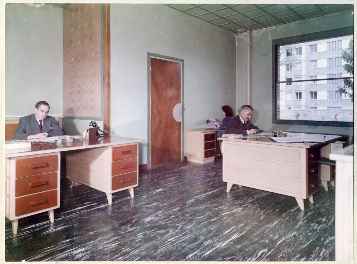 Intérieur du bureau de la comptabilité. Sur le mur à gauche, cloison décorative en PVC réalisée par les Ateliers de Technochimie, les trois bureaux en PVC sont également de fabrication maison. Tirage photographique couleur, 18 x 24 cm, après 1959 (date de construction de l'immeuble visible par la baie vitrée), sans mention d'auteur (collection Ateliers de Technochimie).