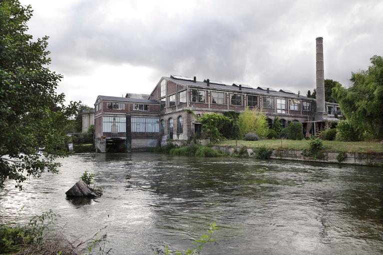 Vue générale des ateliers de la section métal avec le bâtiment de la turbine et la cheminée de l'usine, depuis la rive droite de l'Andelle.