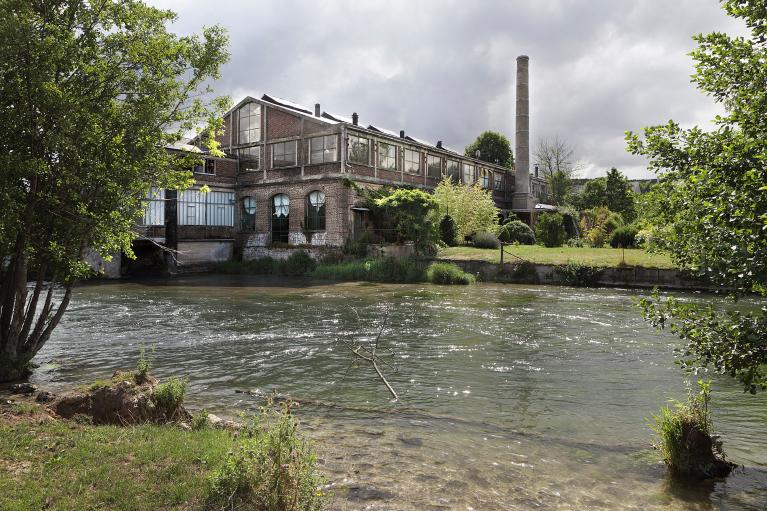 Vue générale d'un des ateliers de la section métal, du bâtiment de la turbine et de la cheminée de l'usine, depuis la rive droite de l'Andelle.