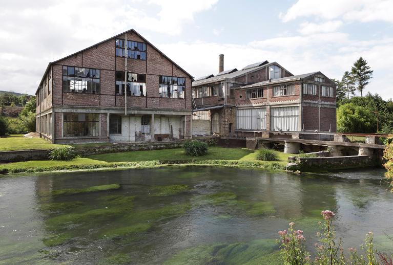 L'usine de jouets Euréka fondée au début du XXe siècle à Douville-sur-Andelle, représentative de l'industrie du bois et du métal du bassin de l'Andelle. ; Vue générale des deux ateliers de la section métal depuis la rive droite de l'Andelle, à gauche l'atelier de polissage, nickelage, conditionnement, à droite l'atelier d'outillage, presse, soudure...) et bâtiment de la turbine. 