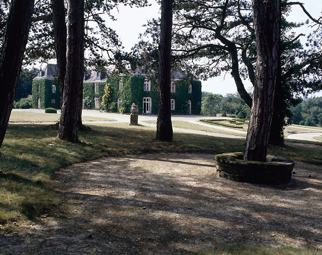 Vue de volume du château prise du sud-est depuis les premiers arbres du parc : au premier plan, banc circulaire autour d'un arbre, puis piédestal avec une potiche. Le château entièrement recouvert de lierre est un élément à part entière du jardin.