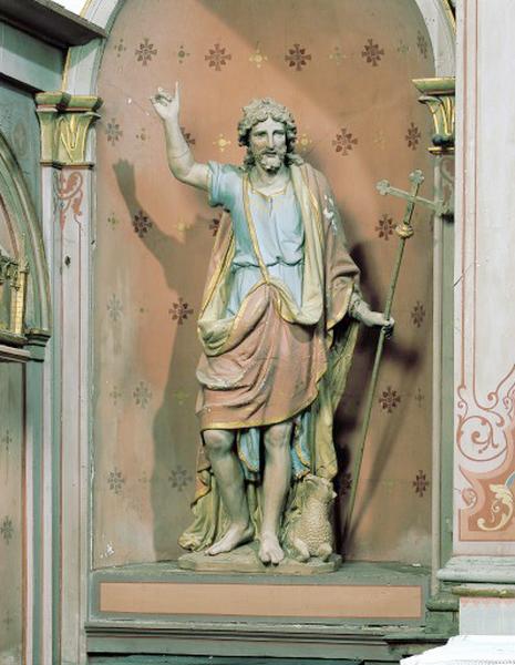 Vue d'ensemble de la statue 17e en terre cuite : le saint est vêtu d'une sorte de toge rouge bordée d'un galon doré et tient de la main gauche une croix en bois peint, sa main droite est levée, à ses pieds est couché un agneau.