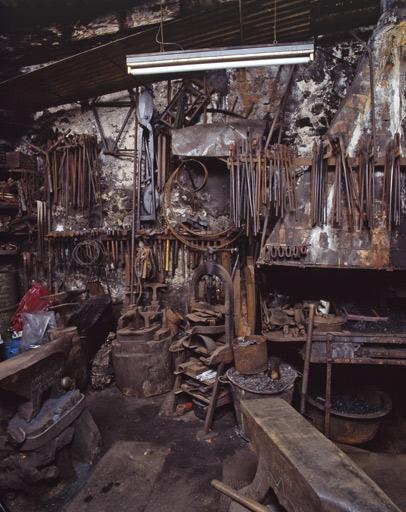 Pinces, goulues, cisailles et marteaux accrochées au mur. Les goulues sont spécifiques à la forge : elles permettent de saisir le lopin (masse de métal) pour le forger. Des bigornes et des enclumes se trouvent au premier plan.