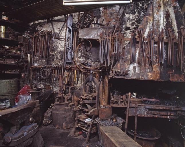 Pinces, goulues, cisailles et marteaux accrochées au mur. Les goulues sont spécifiques à la forge : elles permettent de saisir le lopin (masse de métal) pour le forger. Des bigornes et des enclumes se trouvent au premier plan.