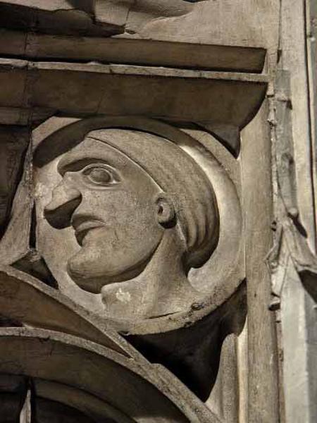 Deuxième travée sud, claire-voie de la troisième section, écoinçon supérieur droit : dans un médaillon circulaire, visage masculin grotesque et de profil portant un bonnet.