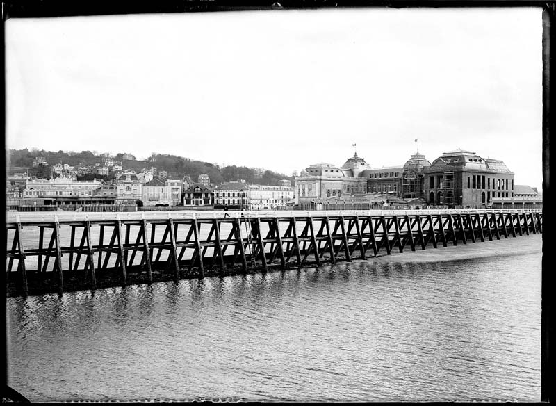 'Chantier de construction. Casino de Trouville. Vue prise de Deauville, avec la jetée au premier plan. Vue des façades nord-ouest et sud-ouest.- Photographie ancienne, n.s., n.d., 1912. Tirage n. et b. d''après plaque de verre gélatino-bromure d''argent (négatif), 13 x 18 cm. (Musée municipal, Villa Montebello, Trouville-sur-Mer. 006.1.96).'