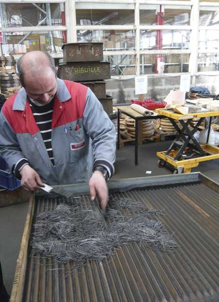 Ouvrier disposant les aiguilles sur un plateau de bois crénelé, afin de les répartir manuellement par vibration en paquets homogènes. Il tient son savoir-faire unique de son père.