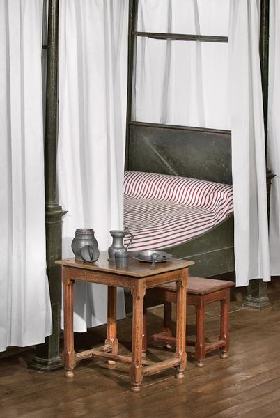 Table et tabouret devant un lit de la salle des femmes.
