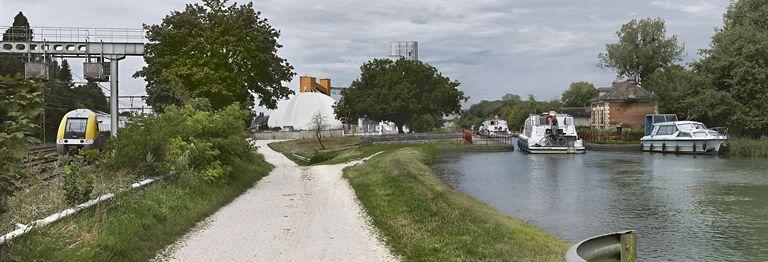 Vue d'ensemble de l'environnement du site d'écluse. De droite à gauche : la maison éclusière, le canal, un silo, la voie ferrée.